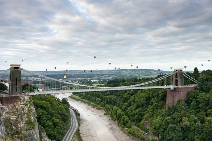 Hot air balloons rising and floating in the air against a cloudy sky, above the city of Bristol and the Clifton Suspension Bridge during the Bristol Balloon Fiesta. River Avon. Credit: Visit Britain